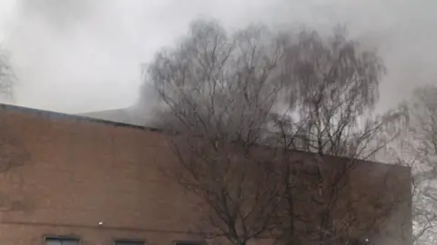 Liam Edwards Smoke billowing into the sky from the top of a large brick building. The building is pictured from the street and has no windows visible. Trees can be seen growing next to it.