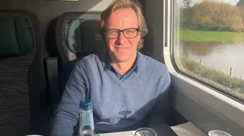 Thomas Kingston in glasses wearing a blue sweater sitting on a train. He is smiling at the camera. Out of the window there are trees and a pond