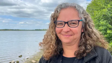 A woman smiles as she stands on the shore of a large lake in front of a green bush. She has curly ginger hair, greying on top, and wears a black jacket, black T-shirt and black-framed glasses. The lake looks grey under a cloudy sky.