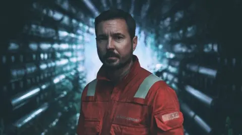 Amazon Prime Video Martin Compston in character on TV show The Rig - he is looking straight into the camera, while wearing a red/orange rig worker's uniform