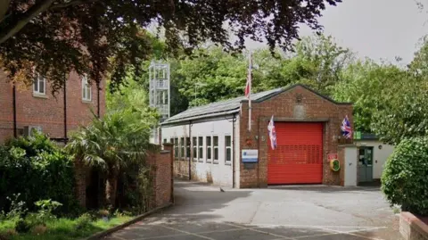 Audlem Fire Station, a brick building with Union Jack flags flying either side of a red garage door. The building is set back on a driveway, with trees surrounding it.