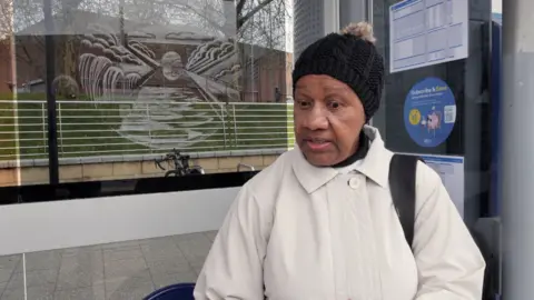 A woman in a white coat and black bobble hat talking at a tram stop