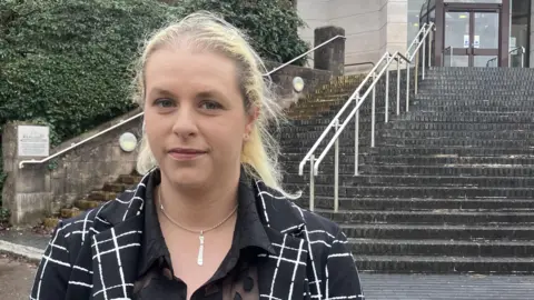 Rachel Serjeant stood outside Newport Crown Court. She is a young woman in her thirties with blonde hair tied in a pony tail and is wearing a black blazer with a white checked pattern. 