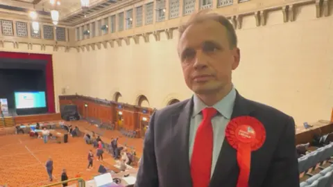 Southampton Itchen MP Darren Paffey stands on the balcony overlooking the election count. He has short fair hair and is wearing a dark suit with a red tie and a red rosette.