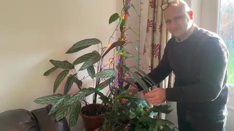 Khaled stands nect to his plants smiling, with his glasses sitting on top of his head. He is wearing a burgundy jumper with a beige collar shirt underneath. His plant has christmas lights wrapped around it.