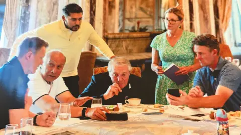Arora Group Four men sitting around a table with one man standing behind and leaning over and a woman standing and holding a large notepad. There are glasses and cups on the table.