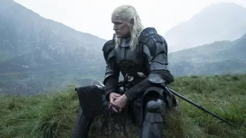 HBO A silver-haired man, sitting on the grass with mountains in the background, wearing a suit of armor