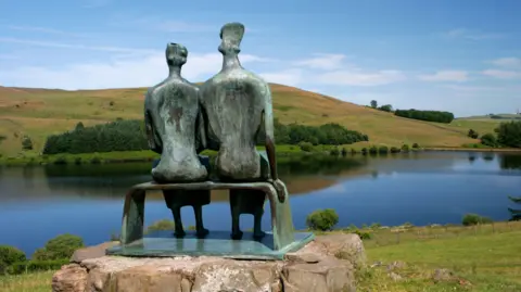 The Henry Moore sculpture King and Queen viewed from behind looking over a reservoir with a tree lined hill in the distance