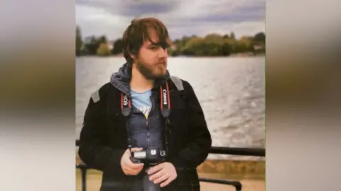 Family photograph Tom James is pictured with a beard and brown hair, standing with a lake or river behind him. He is holding an SLR camera, which is on a strap around his neck.