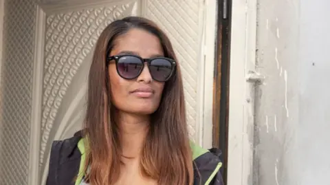 Shamima Begum leaning against a door frame. She has long dyed brown hair and is wearing sunglasses with a light grey top underneath a sweatshirt jacket
