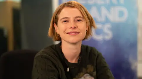 Jessie Buckley, who has short blonde hair and pierced ears with different earrings in. She is wearing a dark green knitted jumper and a bead necklace. She is smiling at the camera and sitting in front of a Desert Island Discs backdrop