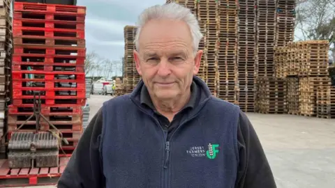 A middle-aged man with slicked silver hair looks into the camera. He's wearing a fleece zip-up gilet, with the 'Jersey Farmers Union' logo and text stitched onto the top left side. Underneath he's wearing a black jumper. He's standing in front off tall piles off red and brown pallet boards.