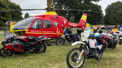 An air ambulance helicopter is stationary in a field with motorbikes parked around it