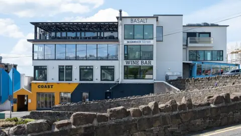 The exterior of the Ramore restaurants building in Portrush. The image shows a tall four storey building with an eatery on each floor. They are marked out by signage, including signs for Coast, Wine Bar, mermaid and Basalt