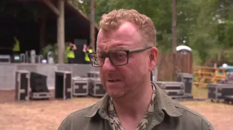 Andy Smith looking to his left. He has short fair hair brushed away from his face and is wearing black glasses and a green jacket. He is standing in front of a stage. A number of boxes containing equipment are on the ground nearby.
