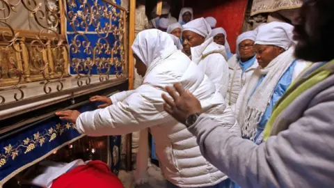 A group of pilgrims wearing light‑coloured clothing gather inside the Grotto at the Church of the Nativity in Bethlehem. Some individuals are reaching forward to touch the ornate, blue‑draped area surrounding the entrance to the small alcove traditionally believed to mark the birthplace of Jesus Christ.