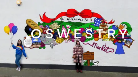 Oswestry Town Council Two women standing in front of a wall with an artist's impression of the colourful mural, featuring the word Oswestry and items that can be bought in the market