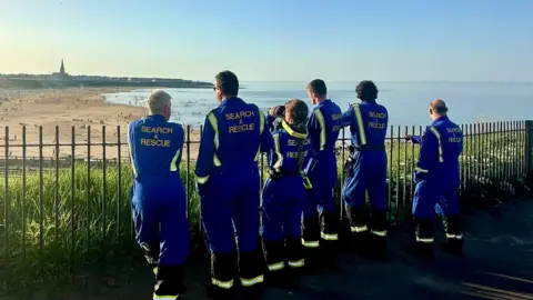 Tynemouth Volunteer Life Brigade A six-strong team of volunteers in blue search and rescue uniform is leaning against a fence watching Tynemouth beach below them. They are pictured from behind and one of them has binoculars trained on the beach.