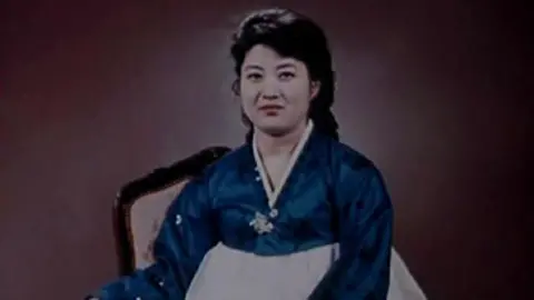 Cheong Seong-chang Ko Yong Hui, probably in her 30s or 40s, wears a blue and white Korean dress and smiles at the camera