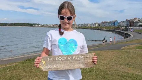 Jonny Smith's daughter holding the wooden sign. It reads The Dock Museum 0.5km next to a small faded yellow arrow. The girl has brown hair and is wearing sunglasses and a white t-shirt with a blue heart on it. She's standing in front of the sea, with houses in the background.