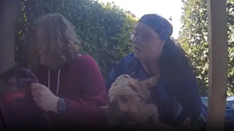 Sam Rousell Two people can be seen on doorbell camera footage trying to open a front door. The woman on the right is holding a dog. The garden path behind them is lined with green hedges. The women appear distressed.