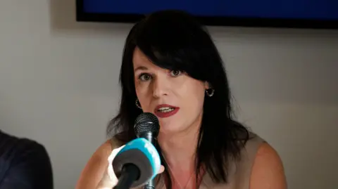 PA Media A woman, sitting in front of a blue microphone. She has a sleeveless cream coloured top on and has long black hair. 