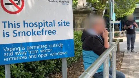 A woman who is wearing a black and light blue jacket and jeans, is sitting smoking outside Hull Royal Infirmary. She is next to a sign which reads 'This hospital site is smoke free.' Her face has been obscured. There is also a man standing a few metres away who is about to light a cigarette. His face has also been blurred. 