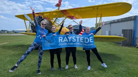 Raystede Centre for Animal Welfare Four women hold a charity banner in front of a yellow plane.