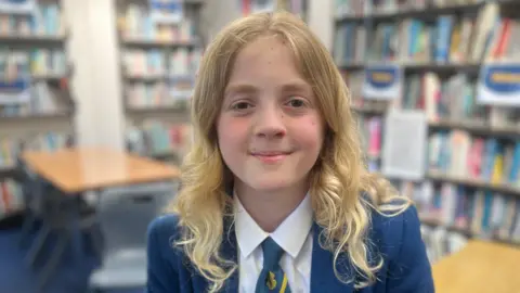 Bea has long curly blonde hair and is wearing a blue blazer, white shirt and blue and yellow tie. She is sitting in a classroom full of books. 
