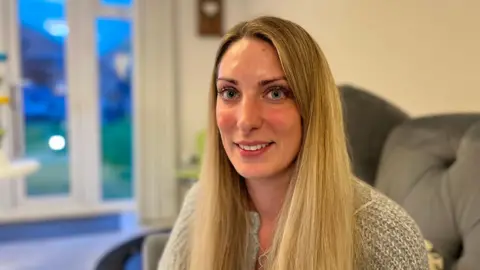 A woman with big blue eyes looks directly at the camera. She has long blonde hair with a knitted top on and is smiling with teeth on show. 