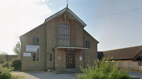 Google Exterior of the church building with a cross on tope of the triangular rood and greenery in front.