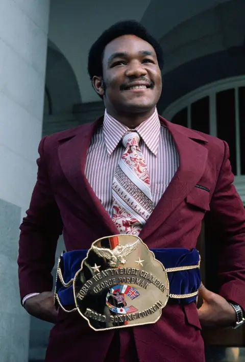 Bettmann via Getty Images Foreman smiling and wearing his heavyweight champion belt