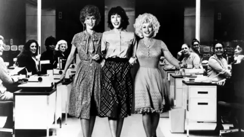 Getty Images/Michael Ochs Archives The three link arms and are pictured walking through the office with desks in the background of this black and white photo. The female typists in the background are all smiling and looking at the three ladies.
