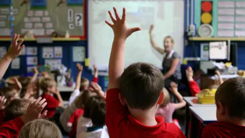 A room full of school children wearing red uniform in a classroom. The boy in the centre of the picture has his hand up to answer a question.