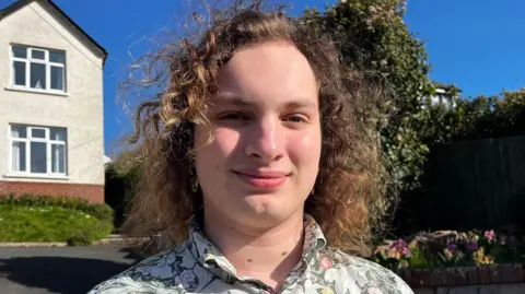 A teenager with curly long hair is standing in front of a house with blue sky in the background. He is wearing a green vintage shirt. 