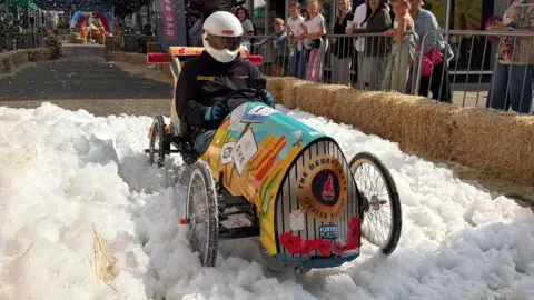 A soapbox car being driven through foam by a person in a motorsports helmet with a visor. The car is painted brightly and reads 'The Desert Rats' on the front. People are standing behind metal crash barriers watching the car drive past.