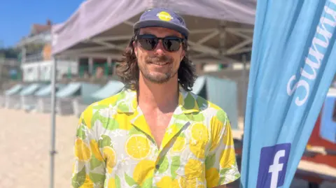 Max Linney, co-director of Sunnyside Deckchairs is stood on the beach in sunglasses and a lemon-pattern shirt. He is stood beside a blue banner and a canopy, with deck chairs below it.