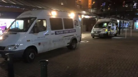 Telford Street Pastors A white van that has Telford Street Pastors written on the side, parked up along a high street. Behind it a police van is also parked