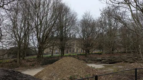 David Spereall/BBC An 18th-century country house, pictured behind a cluster of leafless trees and a mound of rubble.