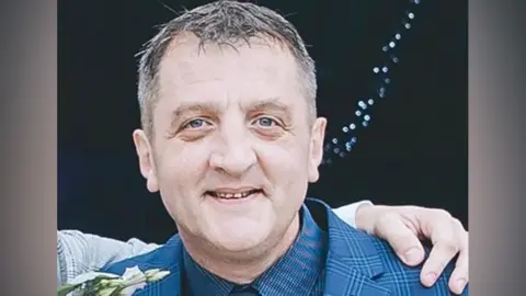 David Dosa: a man with greying hair wearing a blue-patterned jacket smiling. He has a flower in his right lapel and someone's arm is wrapped around his shoulders.
