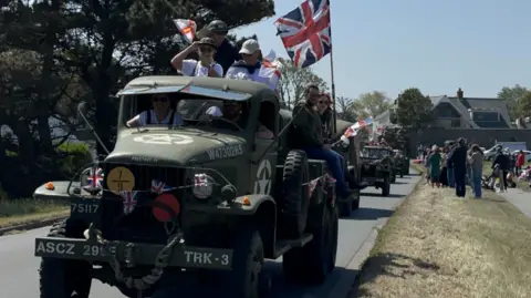 The picture shows a line of vintage military vehicles driving along a road during a parade. The vehicle at the front is an old military truck painted in green with white markings and symbols on the bonnet and sides. The truck is decorated with small flags, including a large Union flag flying from a pole above the cab. Several people are riding in and on the truck, holding flags and waving towards the crowd.
