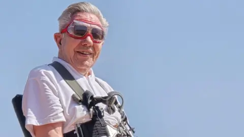 A man with grey hair and wearing a white t-shirt sitting in a chair. He is wearing flying goggles and a black and khaki harness. There is blue sky behind him.