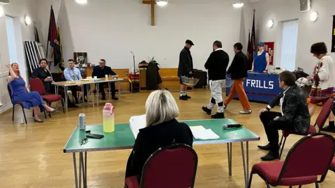 A group of people take part in rehearsals for a play in a room. Some are sat on chairs, others are walking around.
