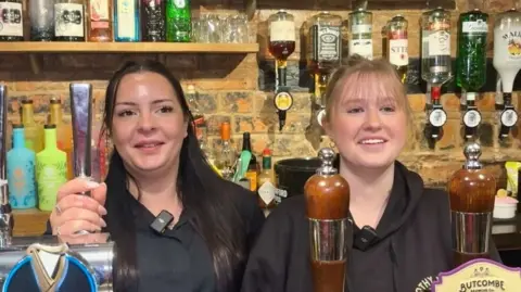 Two women are behind a bar, smiling, They can be seen from the chest up and there are bottles of drinks and optics behind them. The woman on the left has long black hair and a black shirt. The woman on the right has blond hair, tied up and a black shirt.