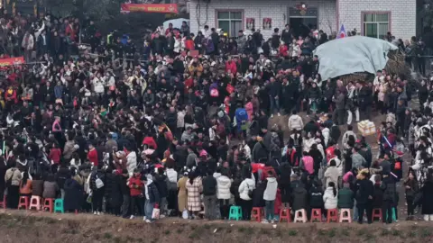 Hundreds of people can be seen standing on the streets outside a white bricked house. The picture is taken from a distance.