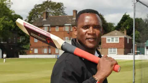 Devon Malcolm at a cricket ground holding a cricket bat over his shoulder and looking at the camera