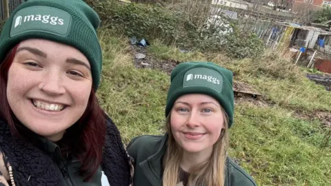 Two women, one with red hair and one with blonde, take a selfie wearing green woolly hats with "Maggs" written on the front. Behind them is a patch of overgrown grass.