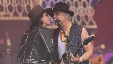 Carl and Pete from The Libertines share a microphone as they perform on a festival stage. 