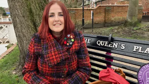 A woman with long red hair and a red and purple chequered coat, is smiling as she sits on a bench outside.