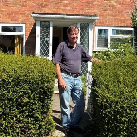 PA Media A man with brown haor stood next to hedges in front of a brick house.